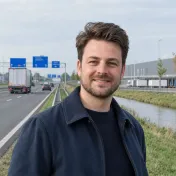 Glimlachende man met donkerblauwe jas naast een snelweg met vrachtwagens en een distributiecentrum